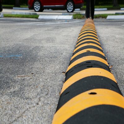 Yellow and Black Striped Speed Bump with Nails Visible in Parking Lot with Diagonal Spaces, White Blocks, Green Grass and Tires of Vehicle Driving by Above in Background